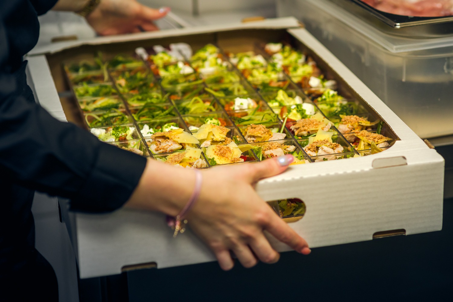 Ręce w czarnej koszuli niosą białe pudełko z cateringiem: porcje sałatek w przezroczystych pojemnikach, widoczne różne kompozycje warzyw, serów i innych dodatków.