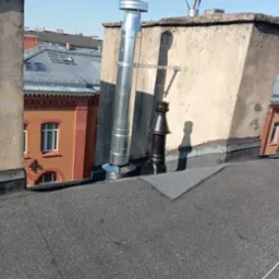 Nowy, srebrny komin wentylacyjny na dachu budynku, zamontowany obok starszego, czarnego komina. W tle budynki z czerwonej cegły i niebieskie niebo.