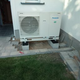 OHSP Sp. z o.o. - Biała pompa ciepła Panasonic R32 zamontowana na betonowych blokach przy ścianie budynku, z widocznymi narzędziami i fragmentem trawnika. Instalacja w toku.