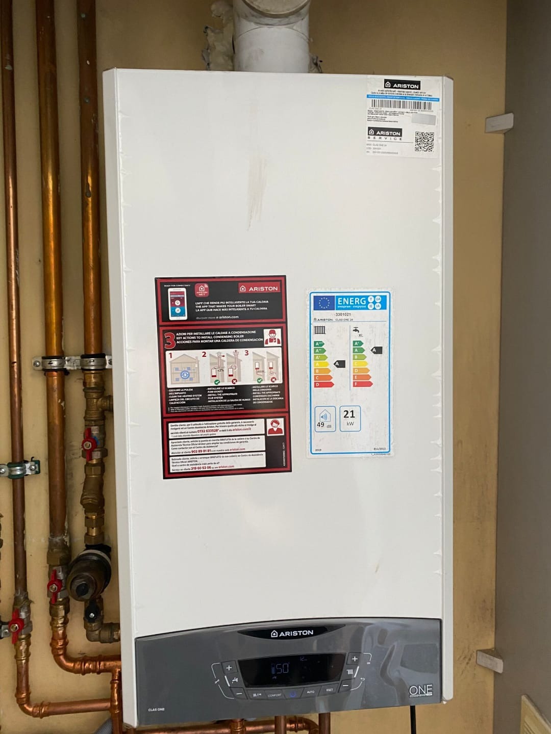Kocioł gazowy Ariston Clas One na ścianie z instalacją hydrauliczną. Widoczne etykiety informacyjne i panel sterowania. Oświetlenie naturalne, lekko z góry.