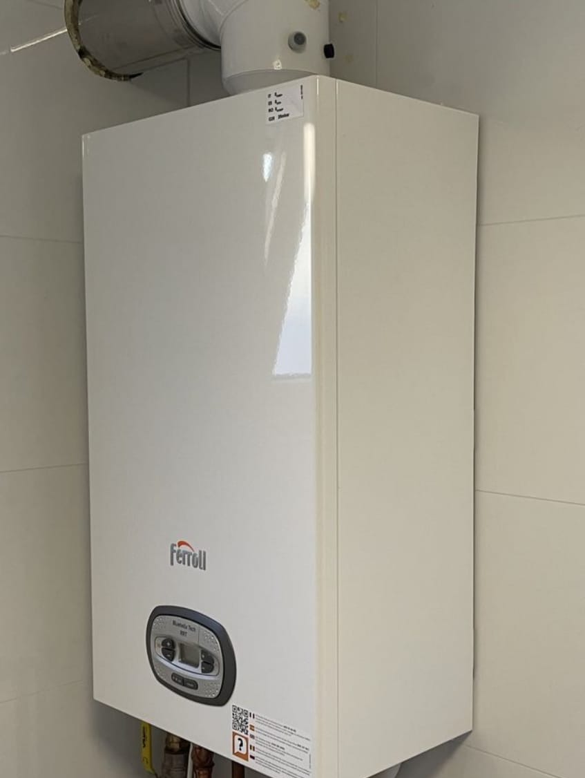 Biały, wiszący piec gazowy Ferroli z podłączonym przewodem kominowym, zamontowany na ścianie wyłożonej płytkami. Widoczne rury instalacji.