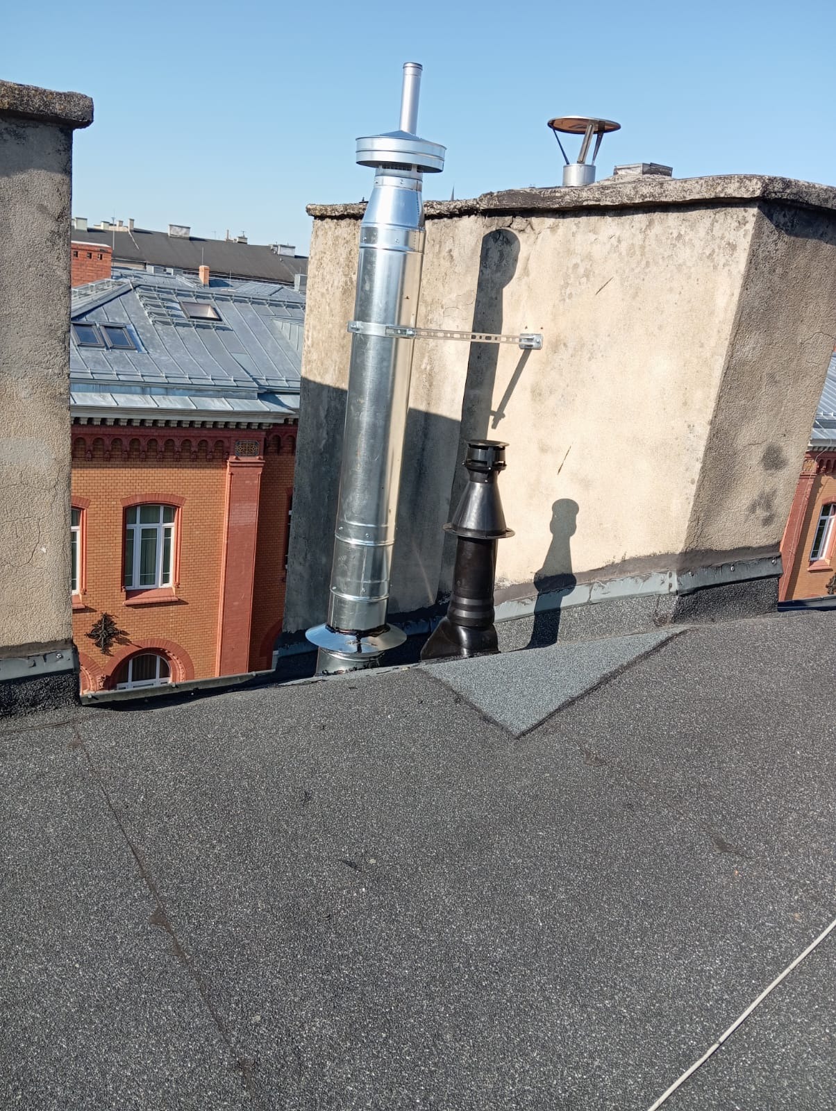 Nowy, srebrny komin wentylacyjny na dachu budynku, zamontowany obok starszego, czarnego komina. W tle budynki z czerwonej cegły i niebieskie niebo.