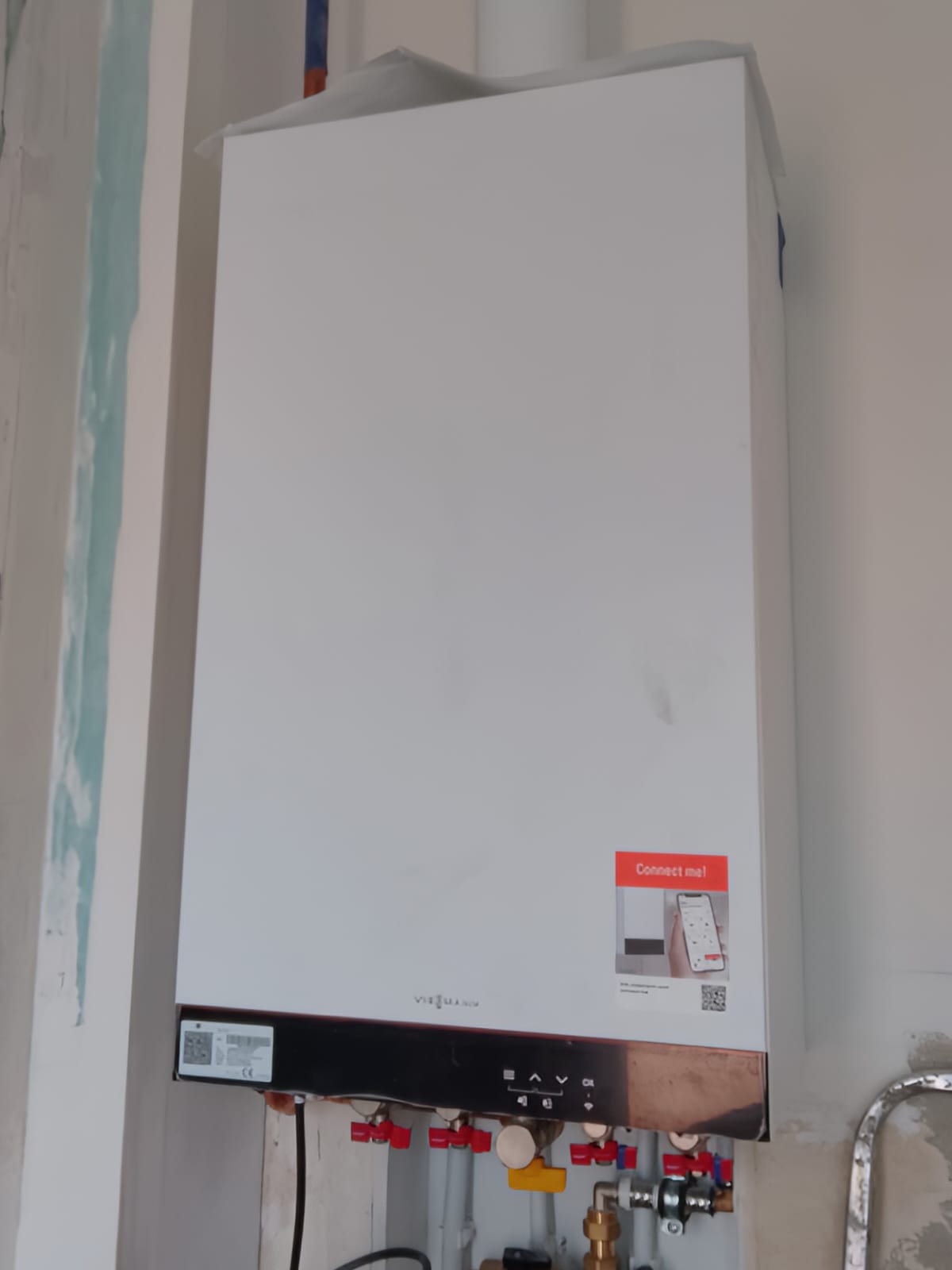 Biały, wiszący kocioł gazowy Viessmann z naklejką 'Connect me!' na ścianie, z widocznymi rurami i zaworami, w trakcie instalacji lub po montażu.