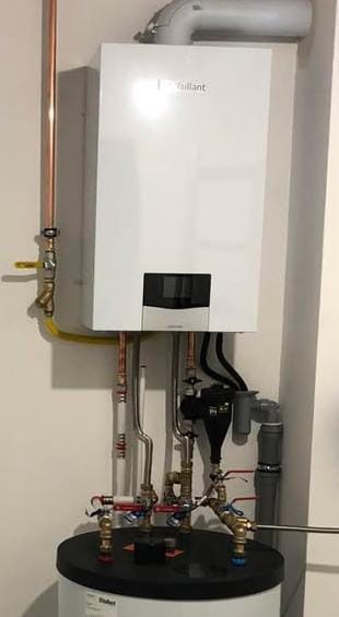 Biały, wiszący piec gazowy marki Vaillant, zamontowany nad zbiornikiem. Widoczne miedziane rury, zawory i przewody. Ujęcie z dołu, instalacja grzewcza w pomieszczeniu.