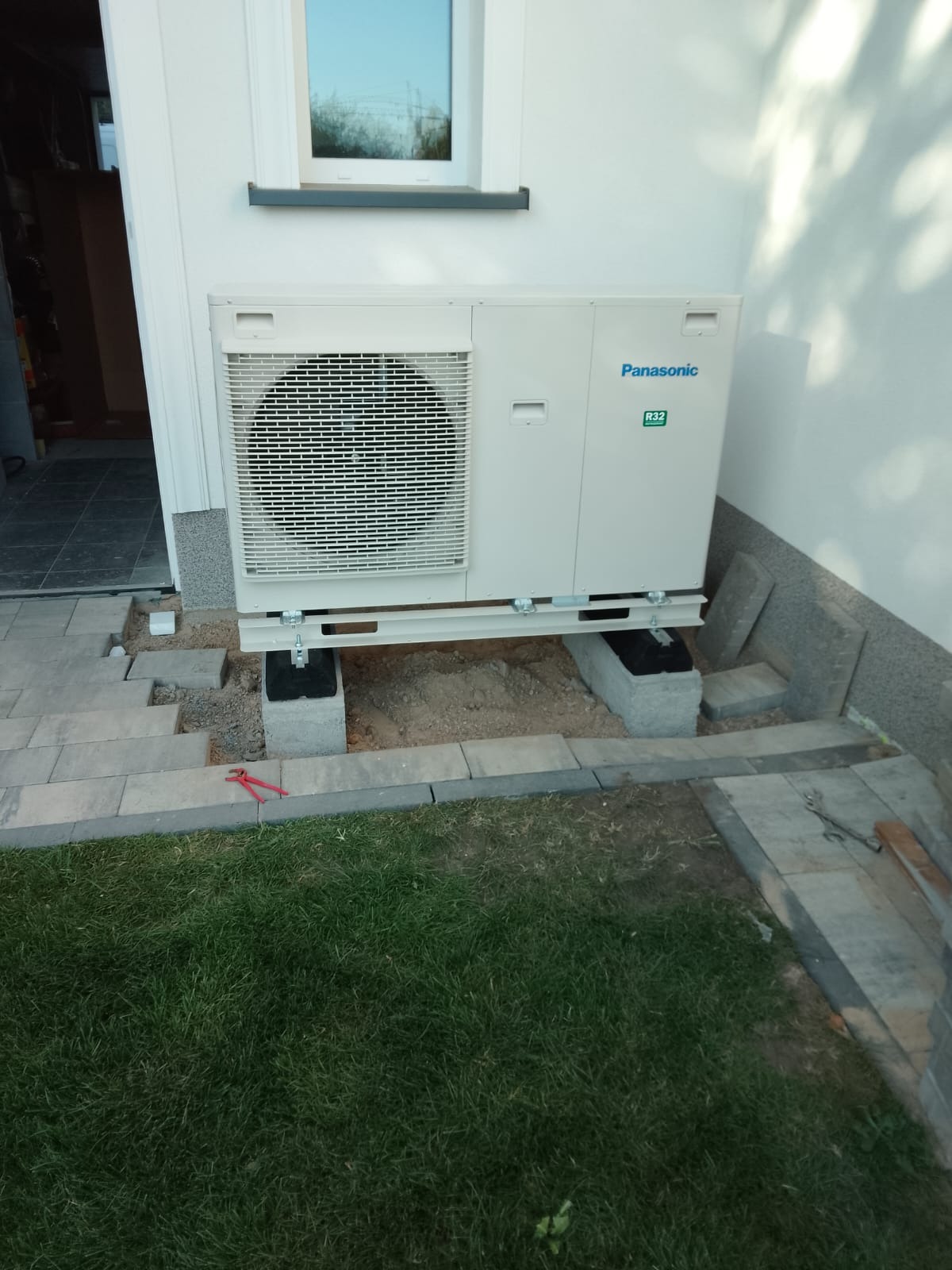 Biała pompa ciepła Panasonic R32 zamontowana na betonowych blokach przy ścianie budynku, z widocznymi narzędziami i fragmentem trawnika. Instalacja w toku.