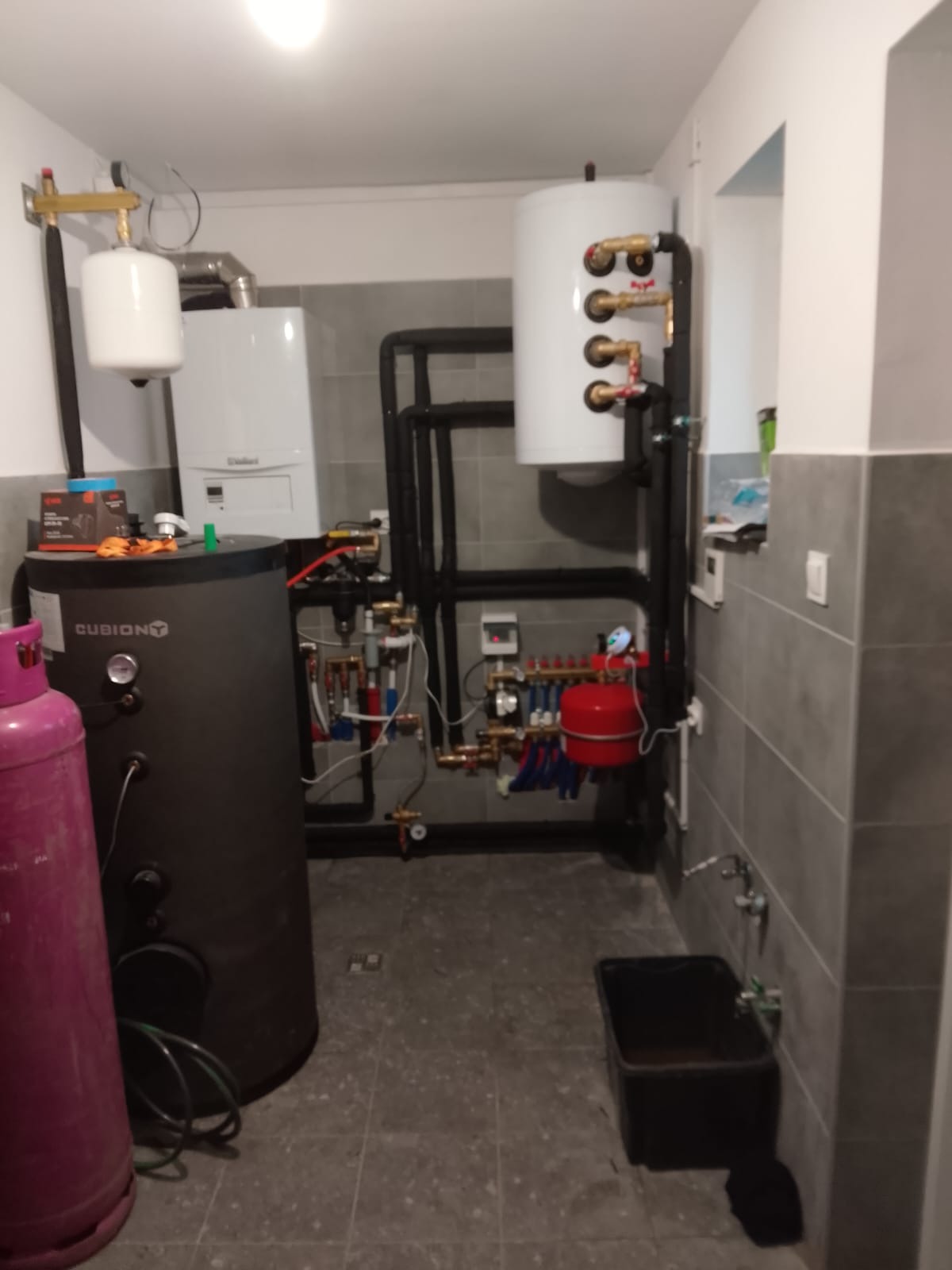Złożona instalacja grzewcza w kotłowni: piec gazowy Vaillant, zbiornik buforowy Cubion, rozdzielacz z pompami i rurami. Szare płytki na ścianach i podłodze.