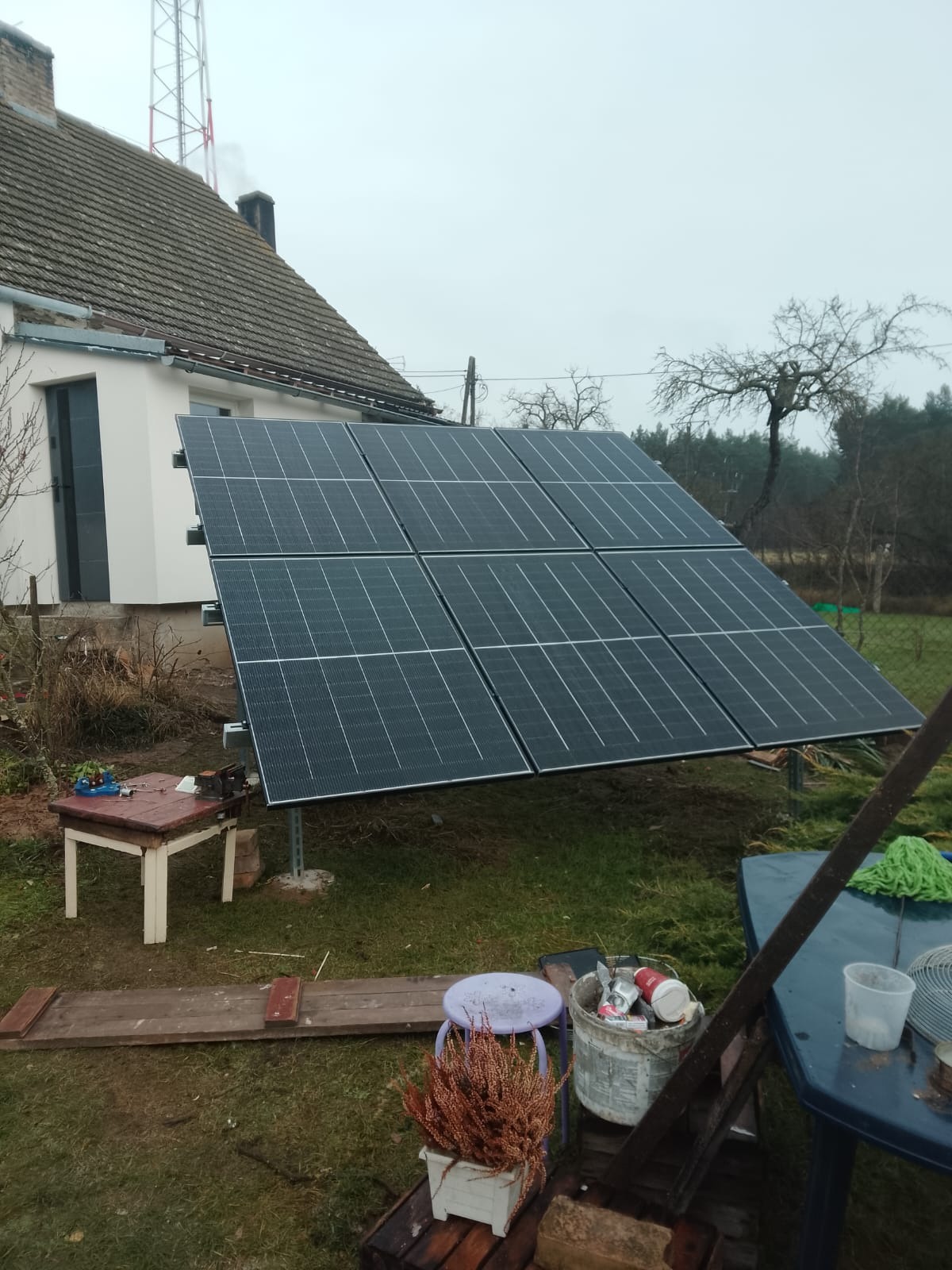 Panel słoneczny na trawniku przed domem, widoczne narzędzia i akcesoria montażowe. Instalacja paneli fotowoltaicznych w pochmurny dzień.