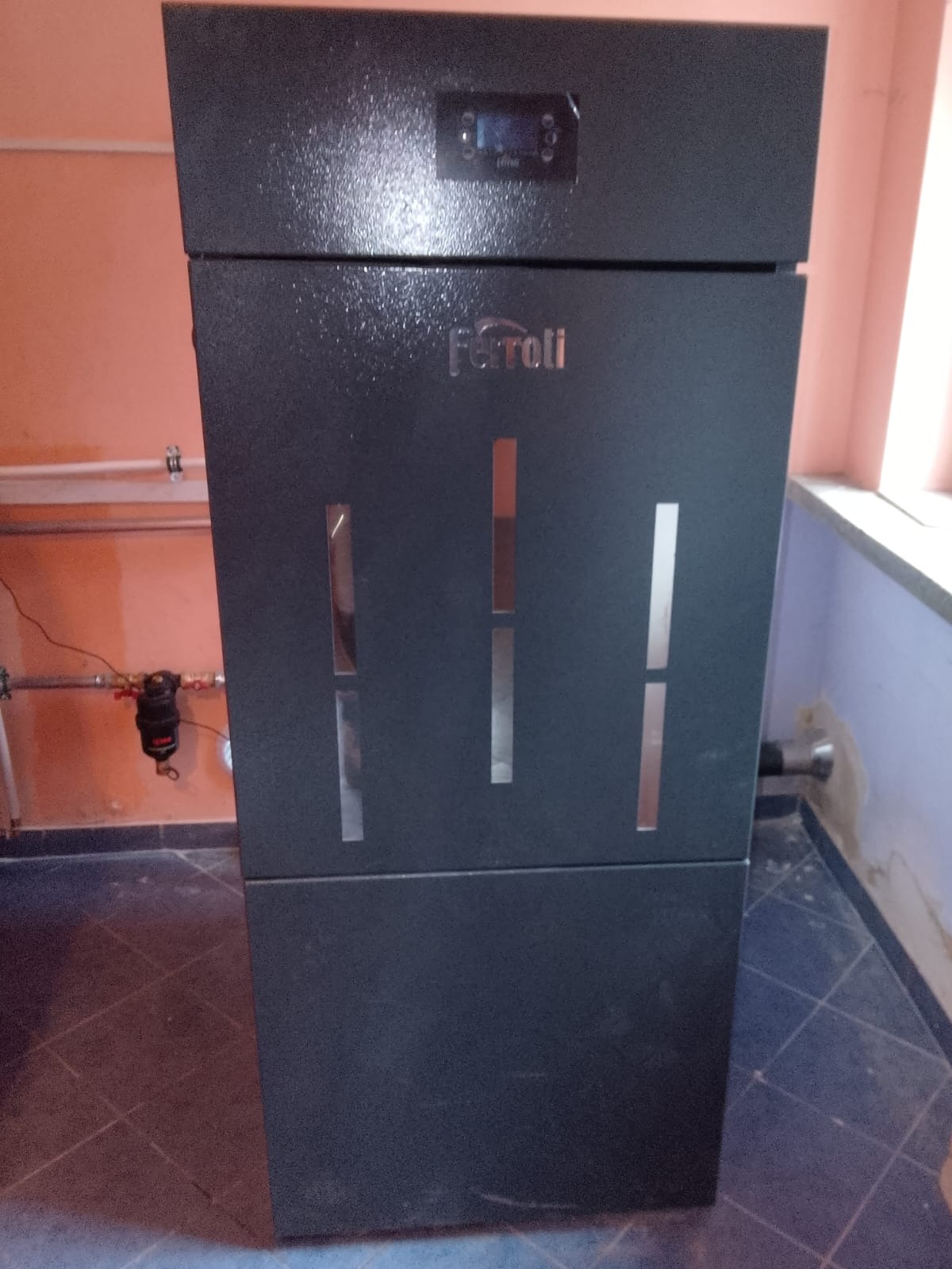 Ciemny piec marki Ferroli z trzema pionowymi szczelinami, ustawiony w rogu pomieszczenia z widocznymi rurami i elementami instalacji grzewczej.