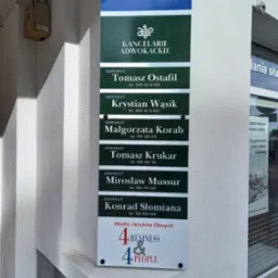 Tablica informacyjna na ścianie budynku z listą adwokatów i ich numerami telefonów, poniżej logo biura tłumaczeń.