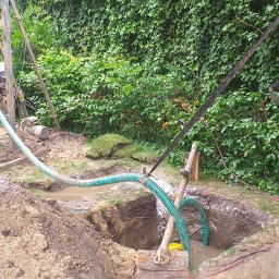 RomBur - Wiercenie otworu w ziemi przy użyciu wiertnicy, widoczne węże doprowadzające wodę, wykopana ziemia, dwie łopaty i bujna roślinność w tle.