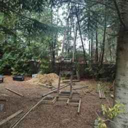 Wiertnica z liną i stertą urobku na zewnątrz, otoczona drzewami i porozrzucanymi rurami.