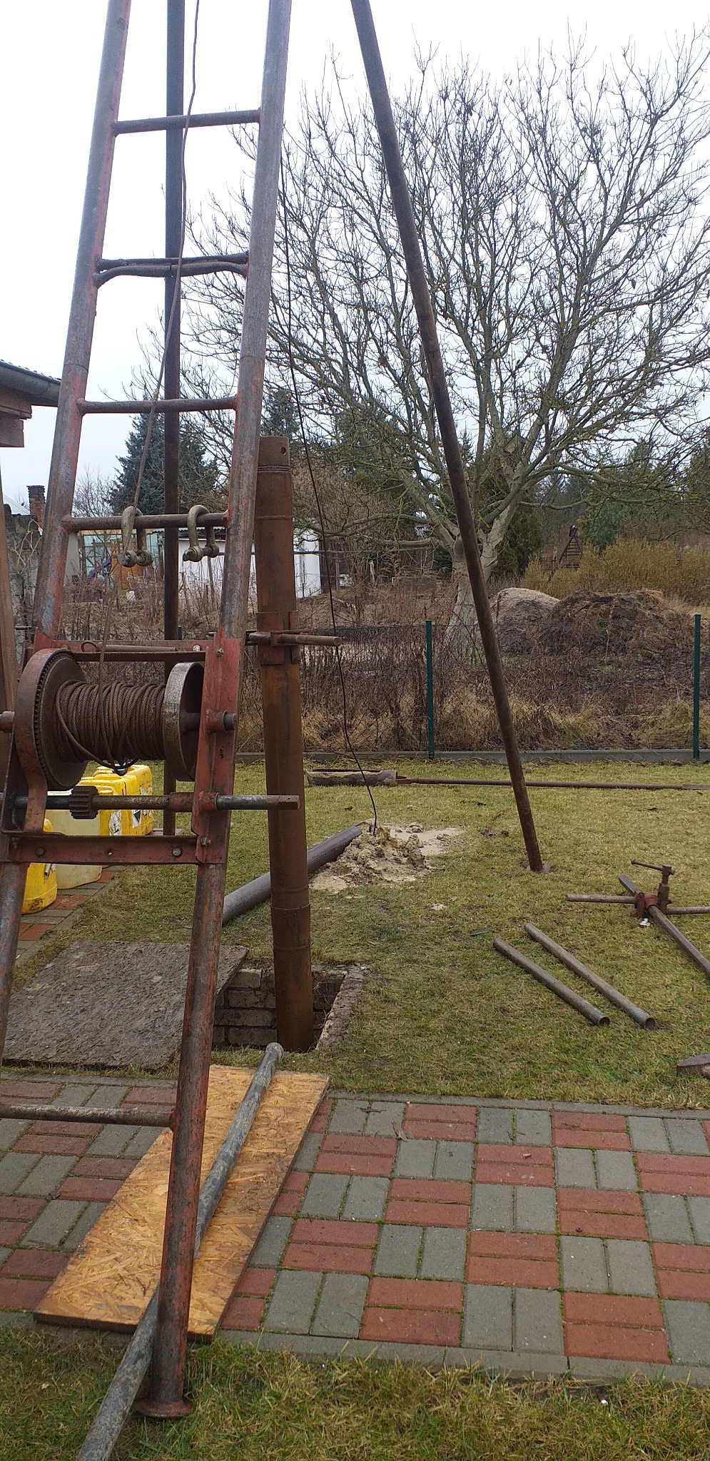 Wiertnica z liną i żurawiem nad otworem w ziemi na trawniku, widoczne rury i narzędzia, w tle drzewo bez liści i ogrodzenie.