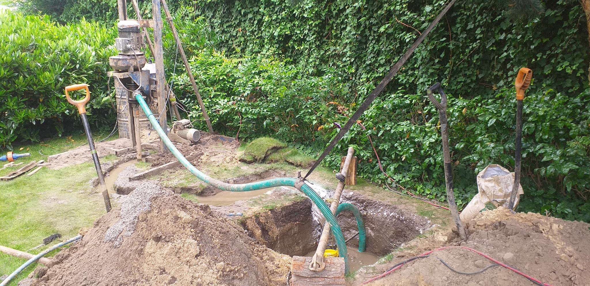 Wiercenie otworu w ziemi przy użyciu wiertnicy, widoczne węże doprowadzające wodę, wykopana ziemia, dwie łopaty i bujna roślinność w tle.