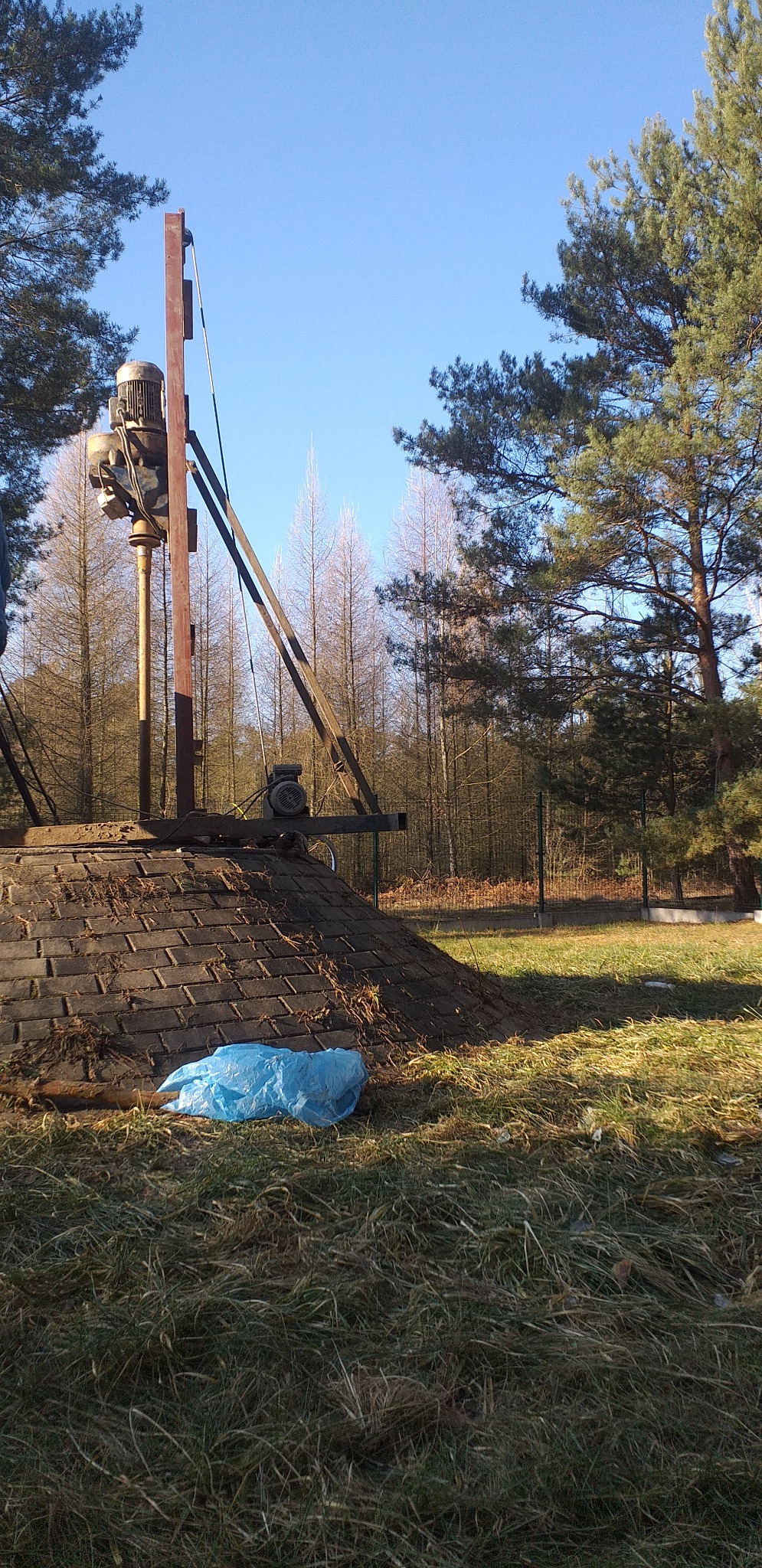 Konstrukcja wiertnicza z silnikiem na szczycie kopca z cegieł, niebieski worek leży na trawie, w tle las i błękitne niebo.