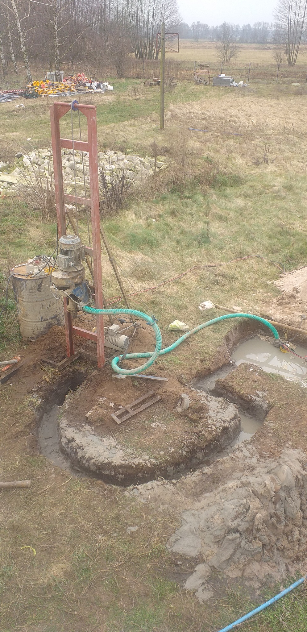 Wiertnica studni głębinowej z silnikiem elektrycznym, woda w wykopie, teren budowy z widocznymi narzędziami i zbiornikiem na wodę.