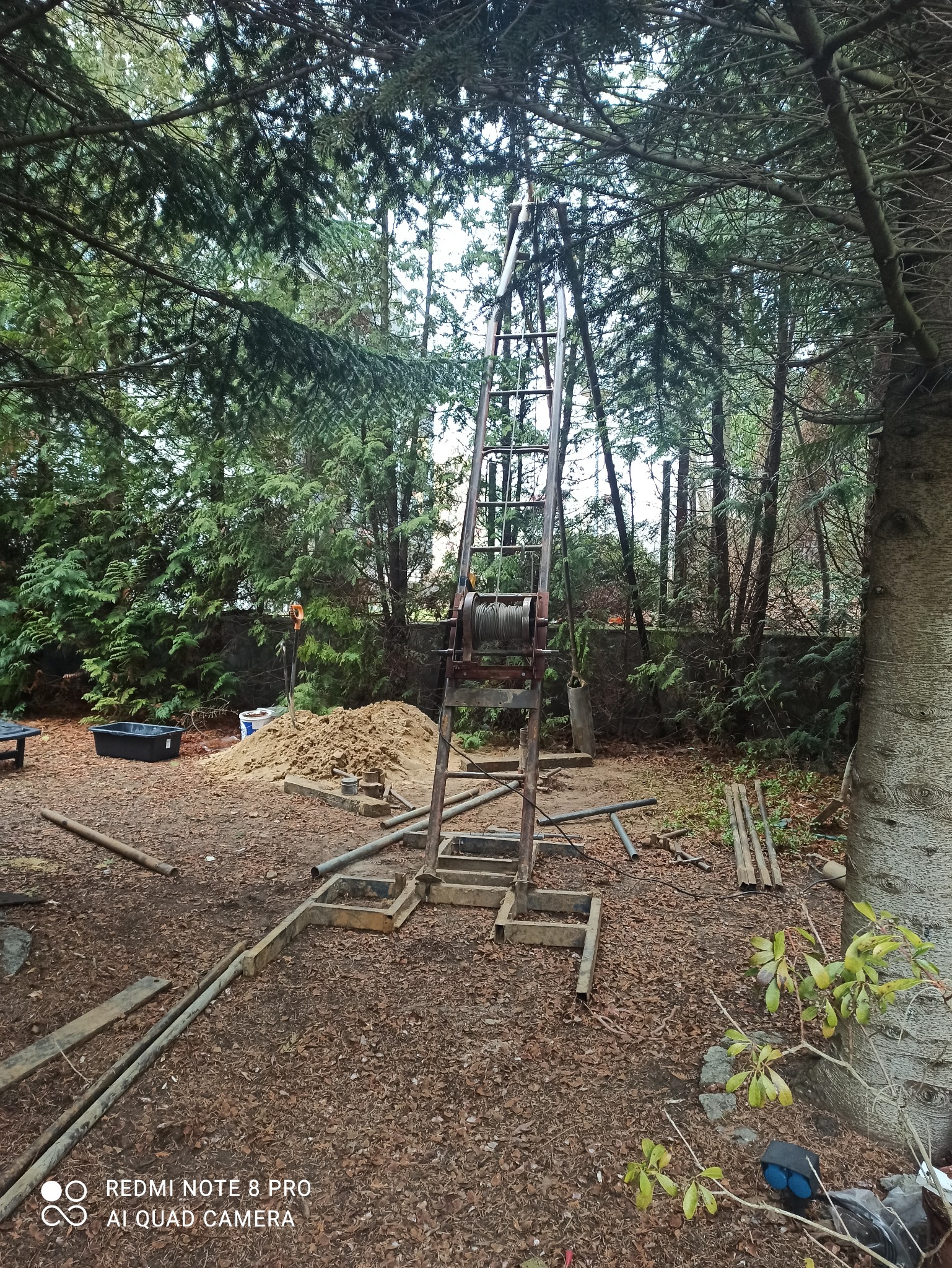 Wiertnica z liną i stertą urobku na zewnątrz, otoczona drzewami i porozrzucanymi rurami.