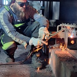 KDS Igor Kosakiewicz - Pracownik w niebieskim kasku i okularach ochronnych spawa grubą metalową konstrukcję, iskry rozświetlają zaciemnione pomieszczenie.