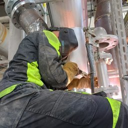 KDS Igor Kosakiewicz - Pracownik w kombinezonie ochronnym z elementami fluorescencyjnymi, w masce spawalniczej i rękawicach spawa metalowe rury z kołnierzami w hali przemysłowej; widoczne iskry spawania.