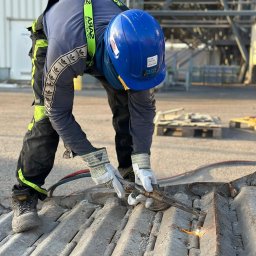 KDS Igor Kosakiewicz - Pracownik w kasku i rękawicach ochronnych używa palnika do cięcia metalowej konstrukcji na zewnątrz, w tle widoczna stalowa konstrukcja hali.