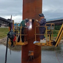 Dwóch spawaczy na podnośnikach koszowych łączy elementy konstrukcji stalowej kolumny na nabrzeżu rzeki, widoczne spawanie i iskry.