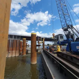 Montaż stalowych pali w wodzie z użyciem dźwigu, widoczny pracownik na podnośniku i fragment mostu w tle.