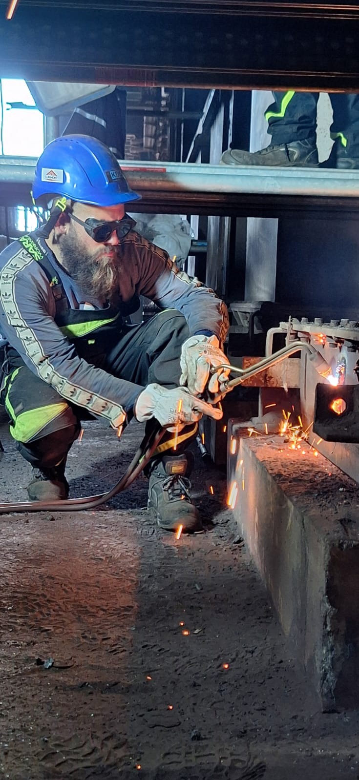 Pracownik w niebieskim kasku i okularach ochronnych spawa grubą metalową konstrukcję, iskry rozświetlają zaciemnione pomieszczenie.