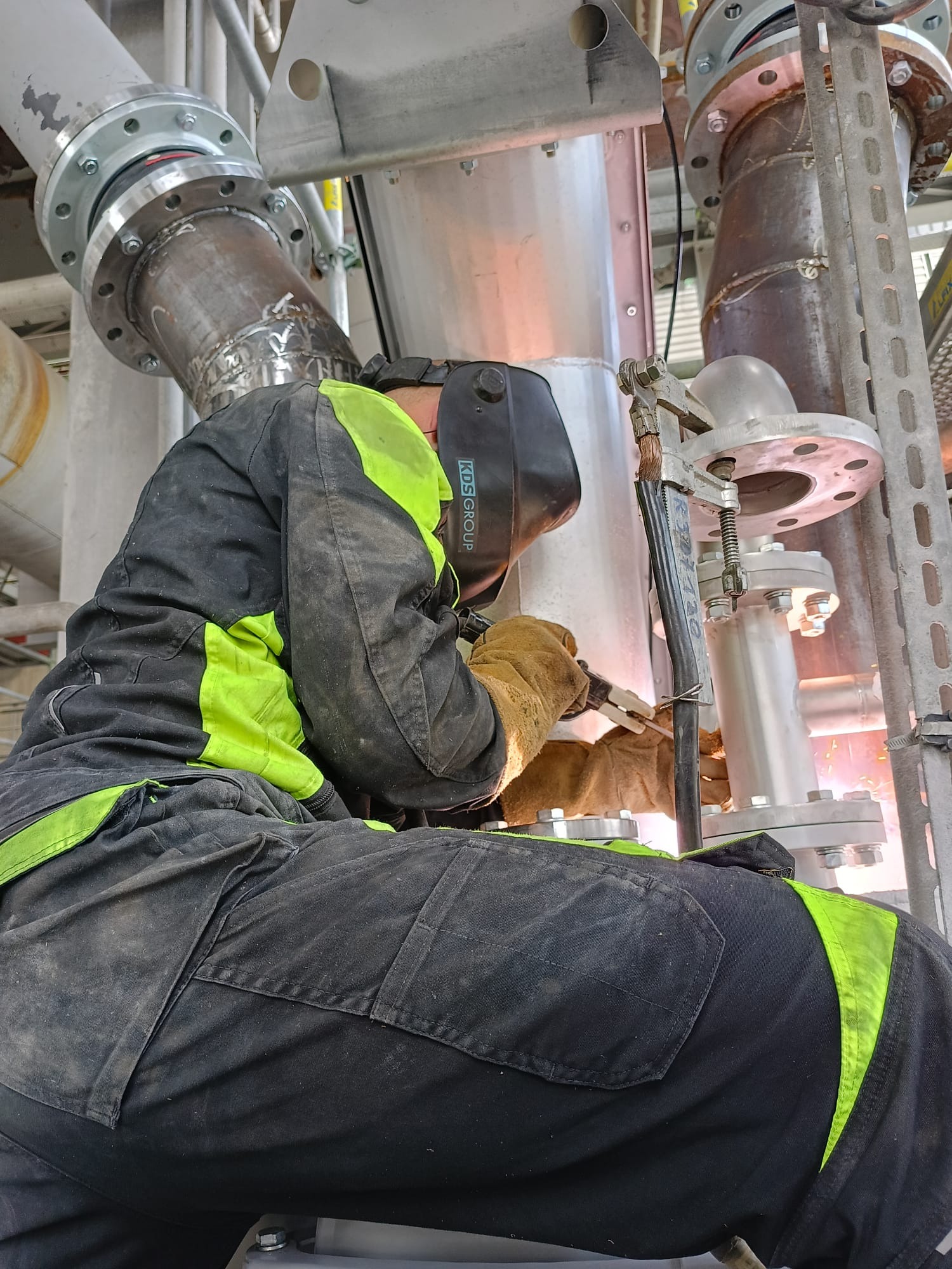 Pracownik w kombinezonie ochronnym z elementami fluorescencyjnymi, w masce spawalniczej i rękawicach spawa metalowe rury z kołnierzami w hali przemysłowej; widoczne iskry spawania.