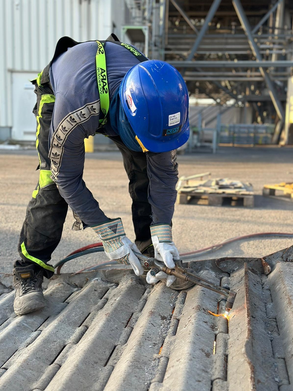 Pracownik w kasku i rękawicach ochronnych używa palnika do cięcia metalowej konstrukcji na zewnątrz, w tle widoczna stalowa konstrukcja hali.