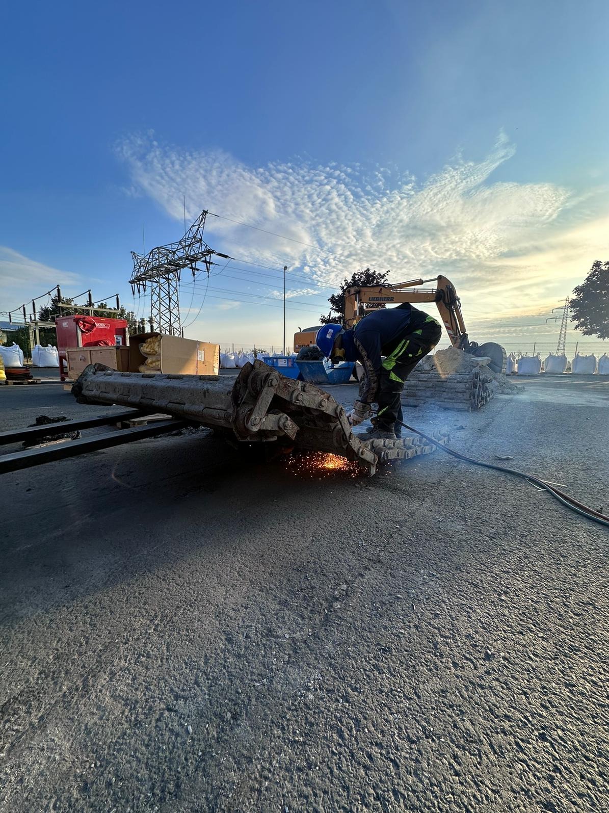 Pracownik w niebieskim kasku spawa metalową konstrukcję łyżki koparki na tle zaparkowanej koparki Liebherr, linii wysokiego napięcia i błękitnego nieba z chmurami.