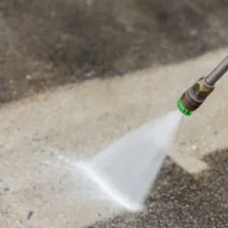 Strumień wody pod ciśnieniem z dyszy lancy czyści fragment betonowej kostki brukowej, pokazując różnicę między czystą a zabrudzoną powierzchnią.