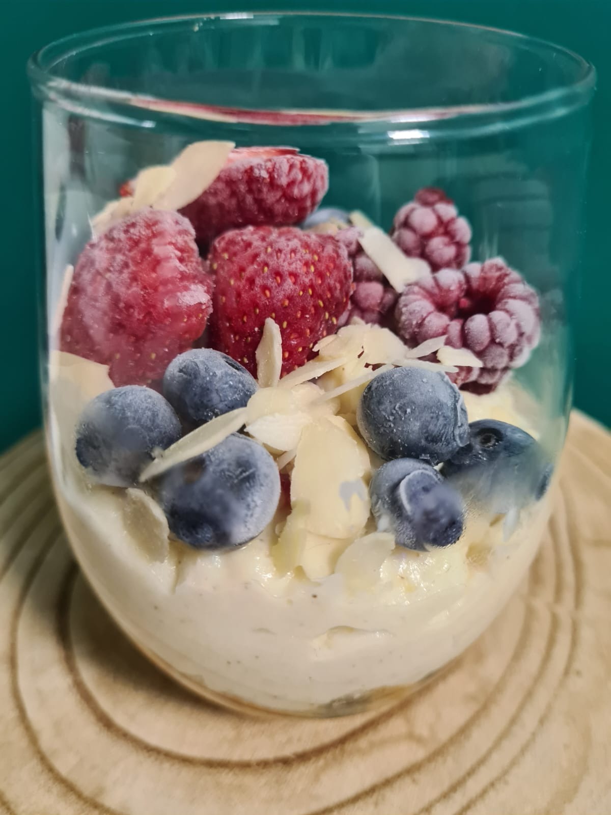 Deser w szklance: kremowy mus z mrożonymi truskawkami, malinami, borówkami i płatkami migdałów na drewnianej podstawce.