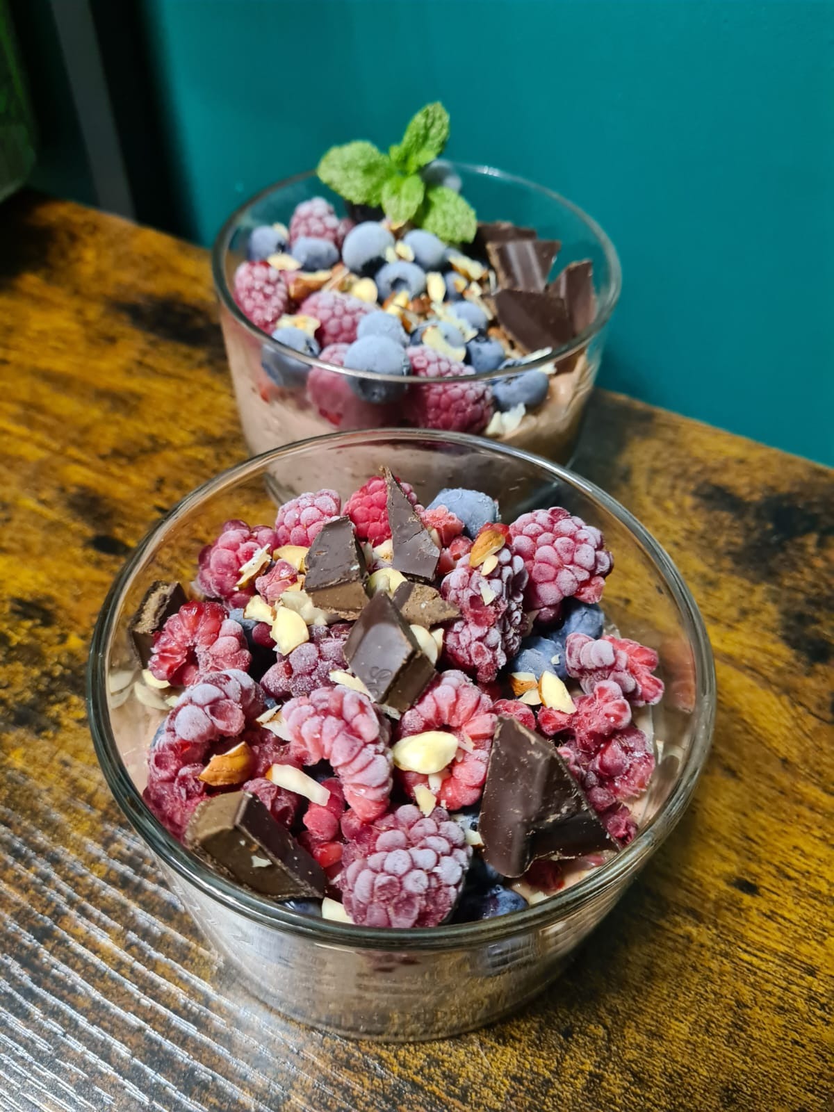 Dwa desery w szklanych miseczkach z mrożonymi malinami, borówkami, kawałkami czekolady i płatkami migdałów, udekorowane listkiem mięty na drewnianym stole i tle w kolorze turkusowym.