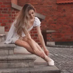Młoda kobieta w białej sukience siedzi na kamiennych schodach, zawiązując tasiemki baletek na tle ceglanej ściany z gotyckim oknem.