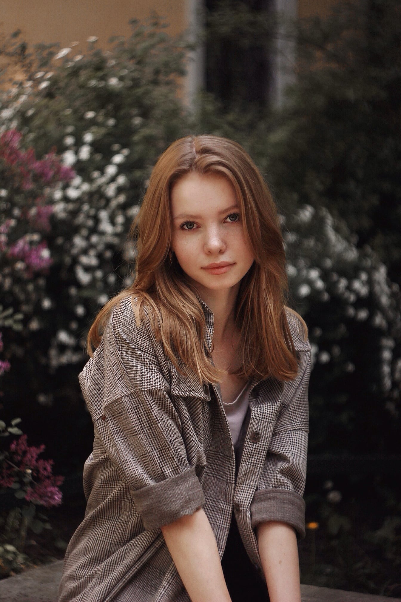 Portret młodej kobiety z rudymi włosami i piegami, ubrana w kraciastą koszulę, na tle zieleni i fioletowych kwiatów.