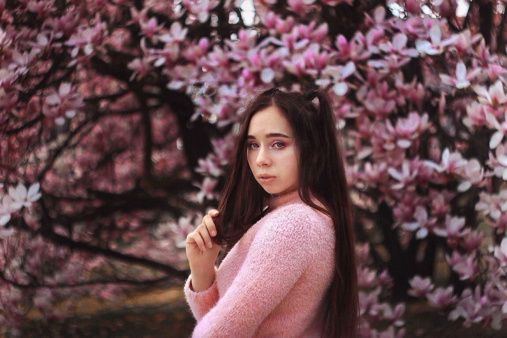 Portret młodej kobiety w różowym swetrze na tle kwitnącej magnolii, z delikatnym makijażem i włosami upiętymi w dwa kucyki.