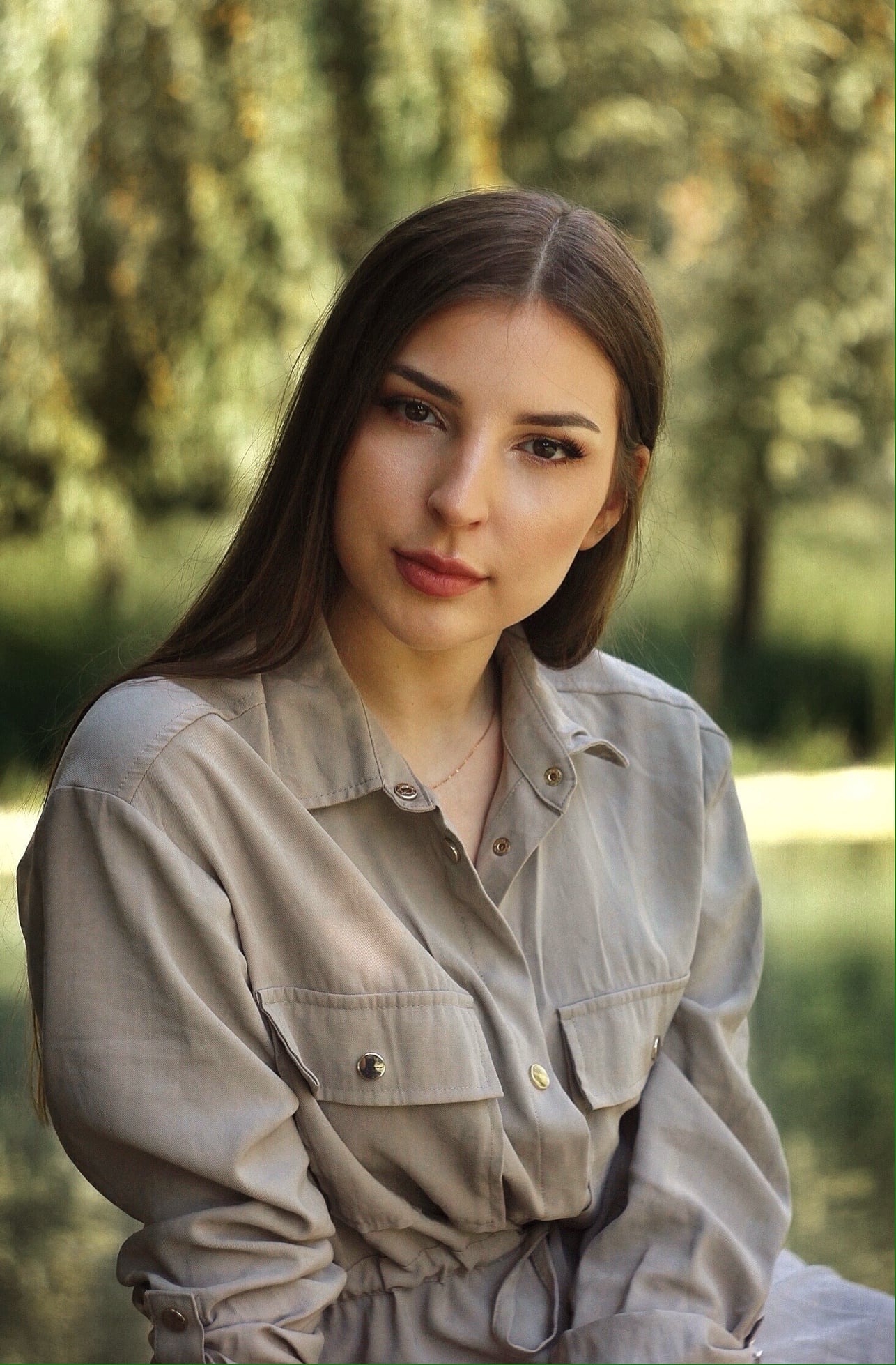 Portret młodej kobiety w beżowym kombinezonie z kieszeniami na tle rozmytej zieleni drzew