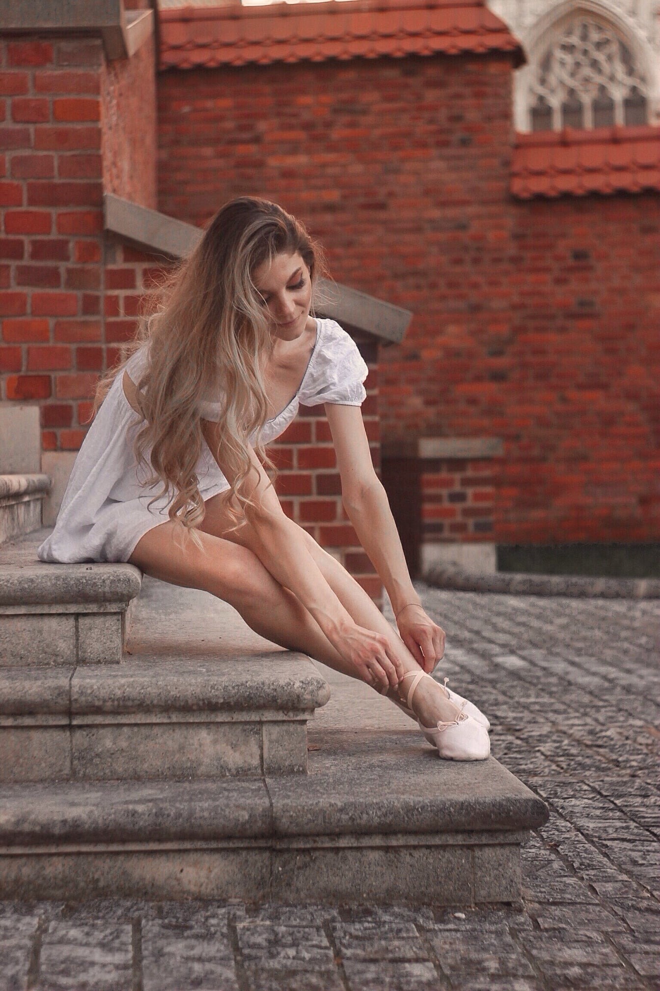 Młoda kobieta w białej sukience siedzi na kamiennych schodach, zawiązując tasiemki baletek na tle ceglanej ściany z gotyckim oknem.
