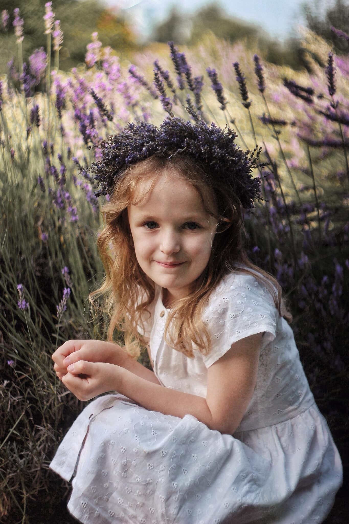 Dziewczynka w białej sukience i wianku z lawendy na głowie, siedząca na tle kwitnącego pola lawendy, z delikatnym uśmiechem.