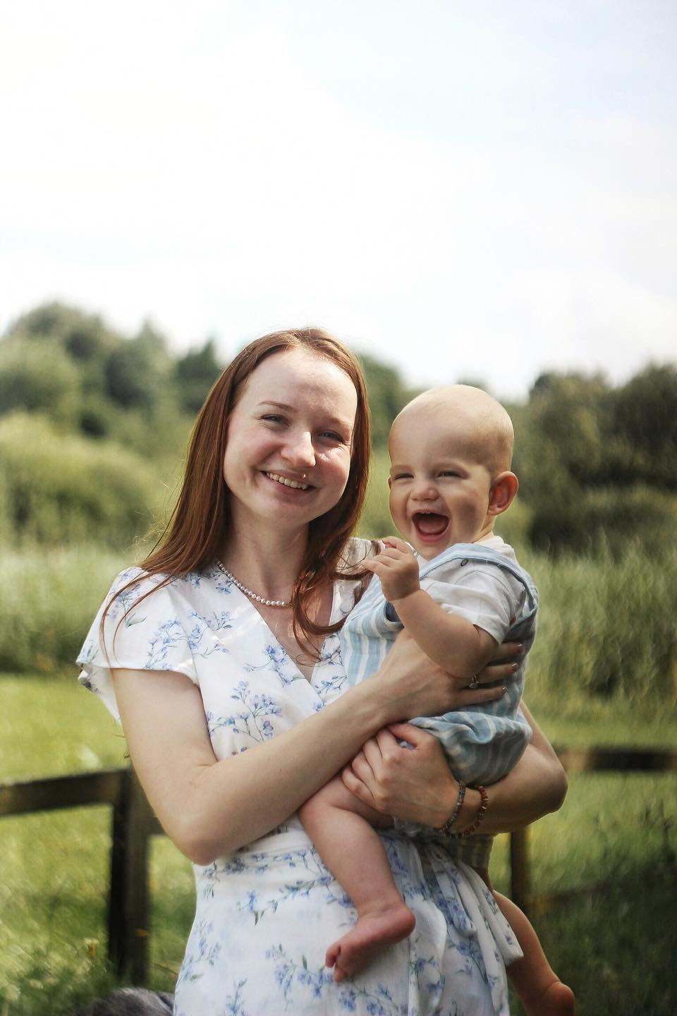 Uśmiechnięta kobieta w kwiecistej sukience trzyma roześmianego niemowlaka w ogrodzie, widoczne drzewa i drewniane ogrodzenie.