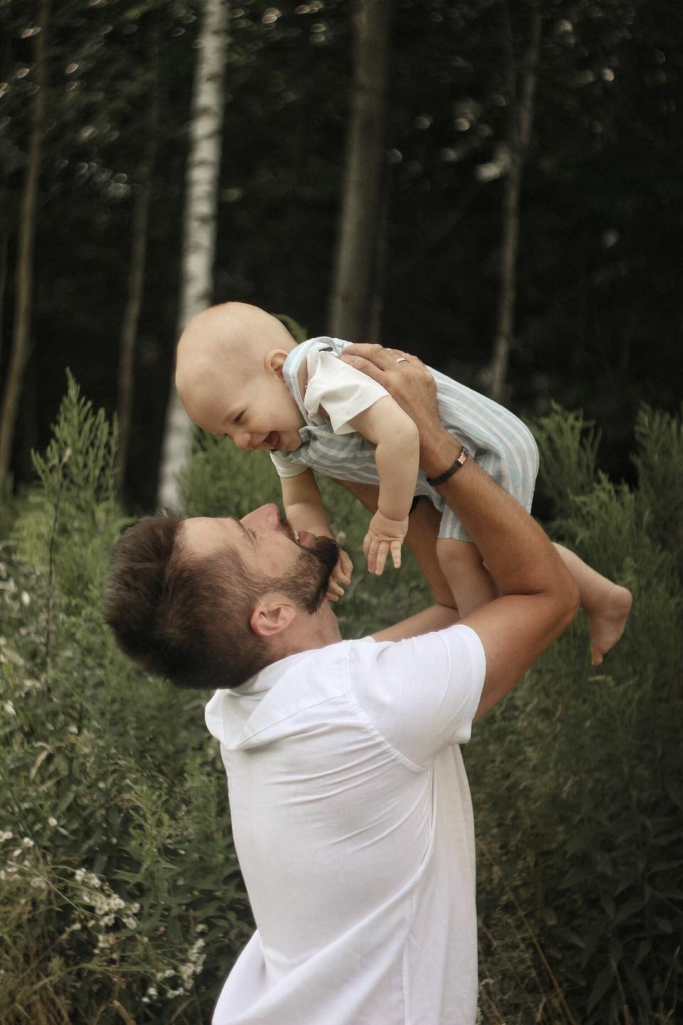 Uśmiechnięty tata w białej koszuli podnosi wysoko roześmiane dziecko w paski na tle zieleni i drzew.