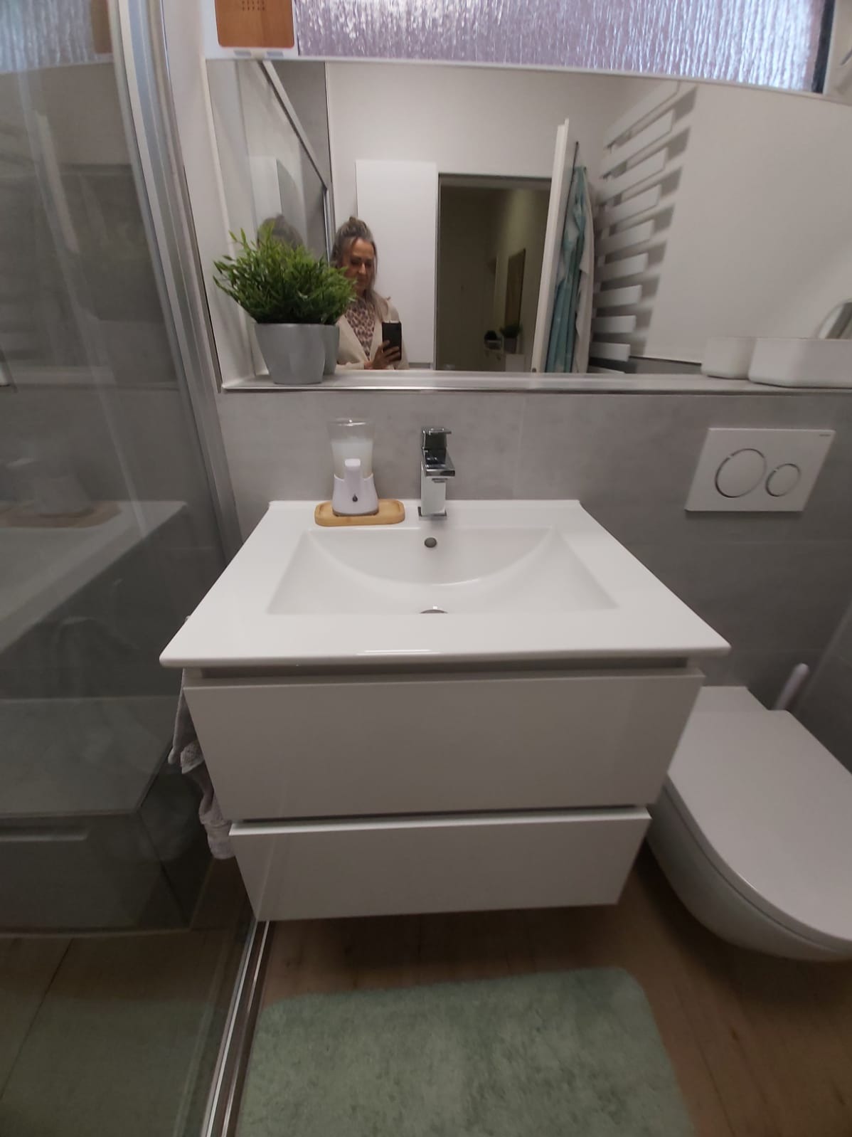 Nowoczesna, jasna łazienka po remoncie: biała umywalka z szafką, szara ściana, toaleta i lustro z odbiciem osoby robiącej zdjęcie.