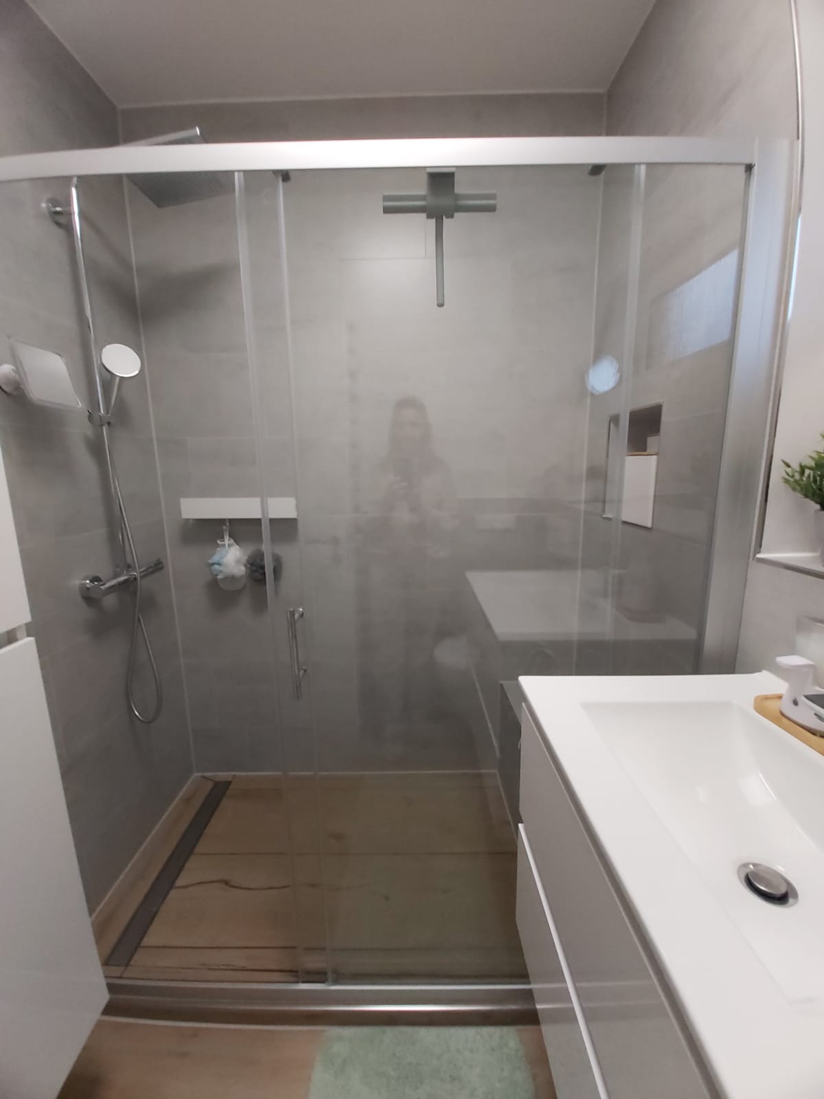 Nowoczesna kabina prysznicowa z szarymi płytkami, drewnopodobną podłogą i minimalistyczną armaturą. Widoczna umywalka z szafką w kolorze białym.