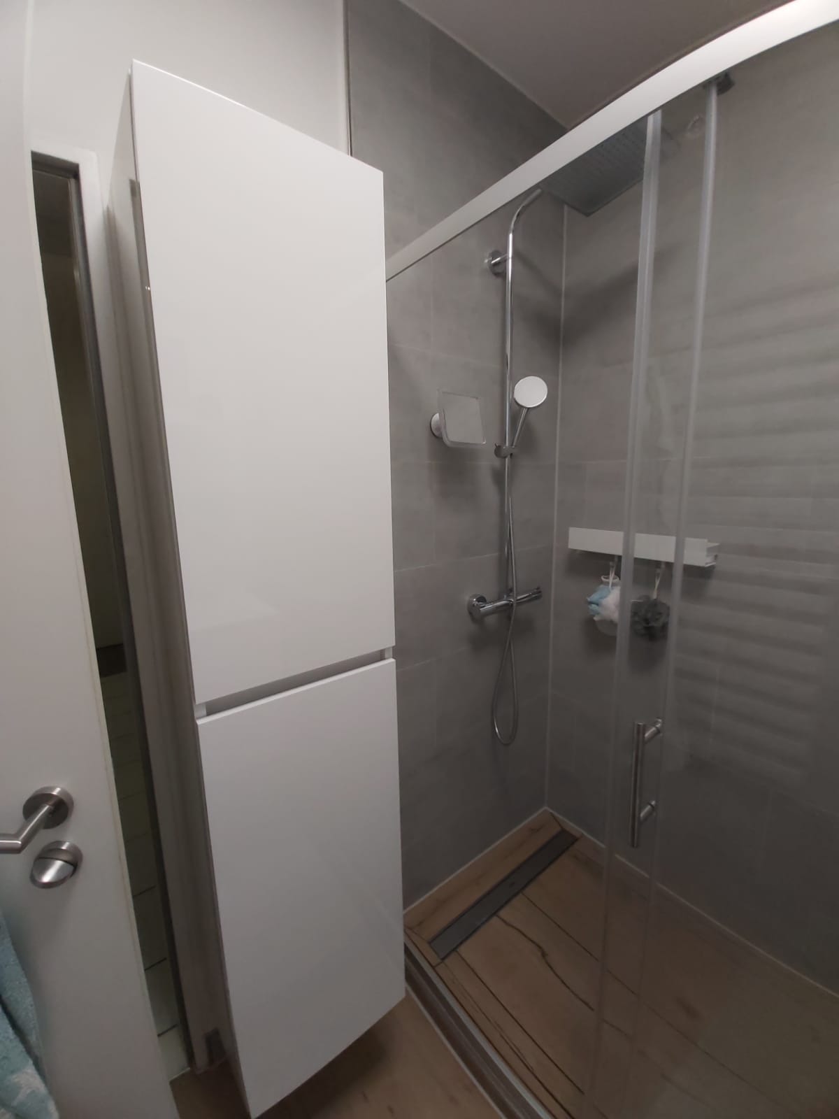 Nowoczesna łazienka z kabiną prysznicową typu walk-in, szarą glazurą i białym, wysokim regałem. Widoczny fragment drzwi i drewnopodobna podłoga.