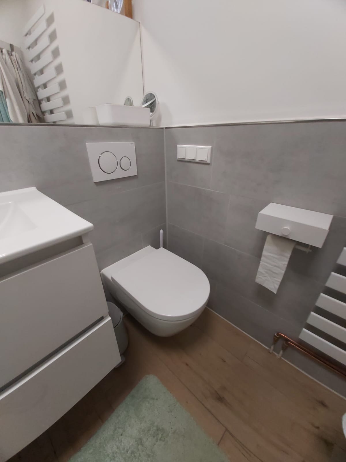 Nowoczesna, minimalistyczna łazienka w szarościach i bieli z drewnianą podłogą. Uwagę przyciąga podwieszana toaleta i grzejnik drabinkowy z miedzianymi elementami.