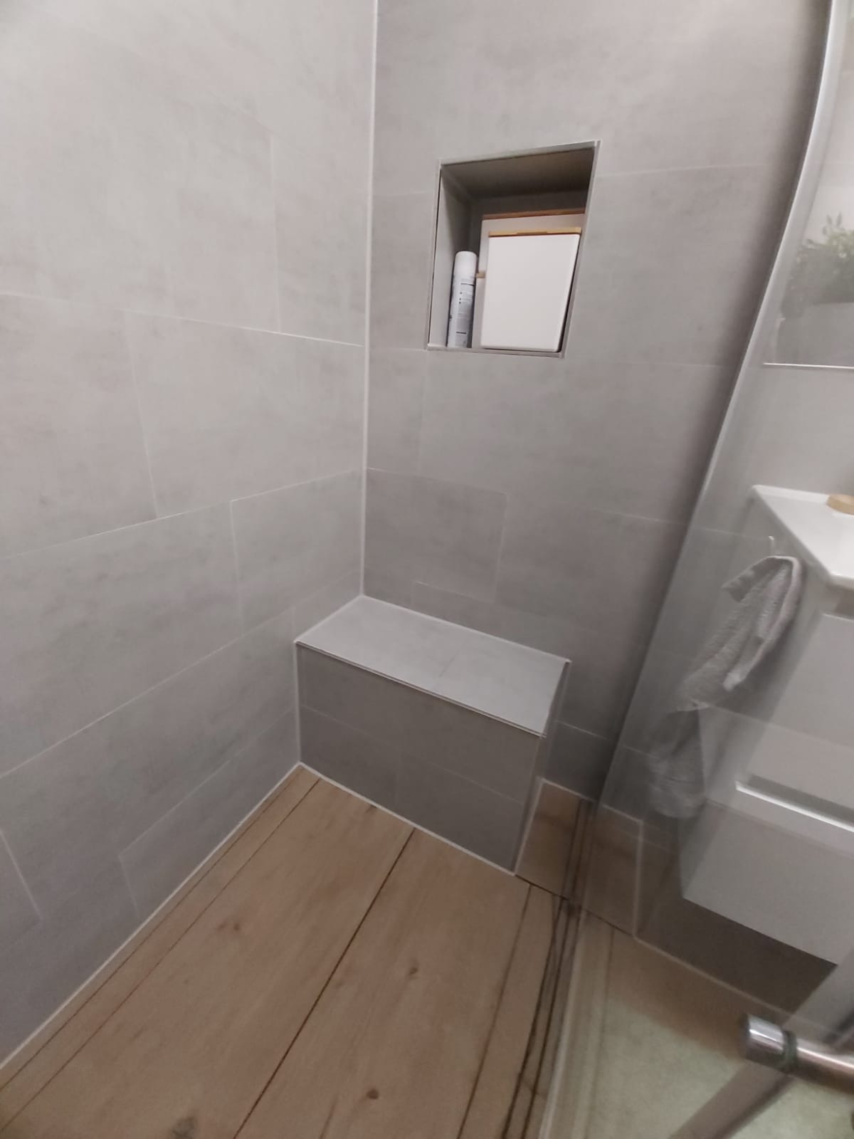 Wykończona łazienka z szarymi płytkami, drewnopodobną podłogą, wnęką w ścianie i zabudowanym siedziskiem. Nowoczesny design z dbałością o detale.