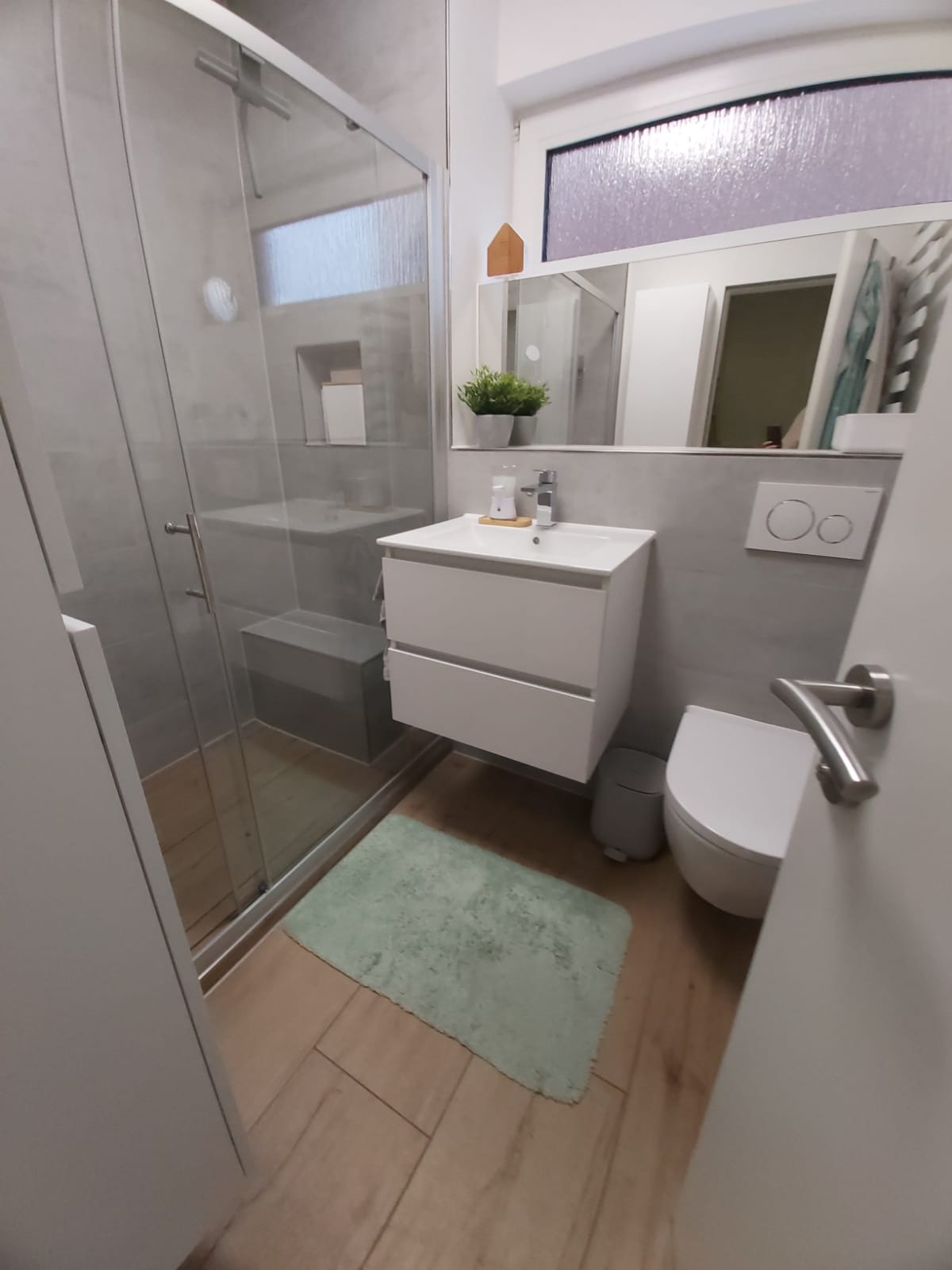Nowoczesna łazienka z kabiną prysznicową, umywalką z szafką, toaletą i lustrem. Jasne płytki, drewnopodobna podłoga i zielony dywanik tworzą przytulną atmosferę.