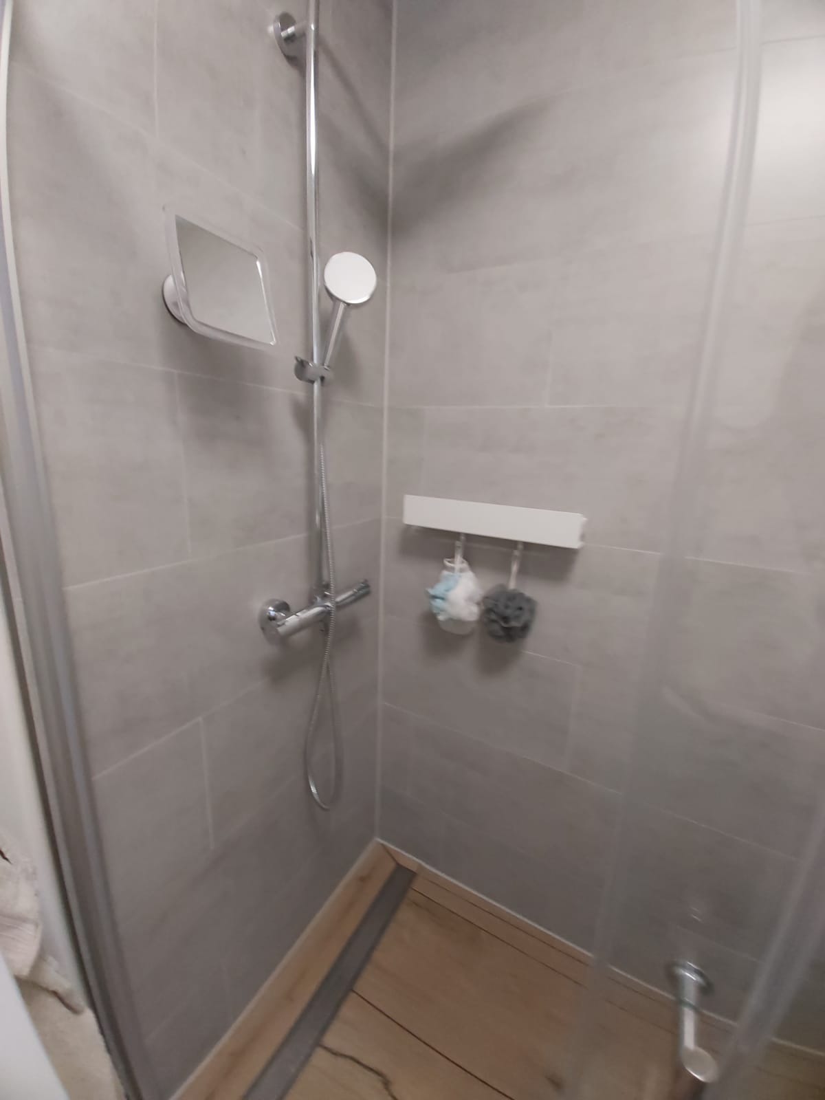 Nowoczesna kabina prysznicowa z szarymi płytkami, deszczownicą, i drewnopodobną podłogą. Na ścianie zamontowane lusterko i półka z akcesoriami kąpielowymi.