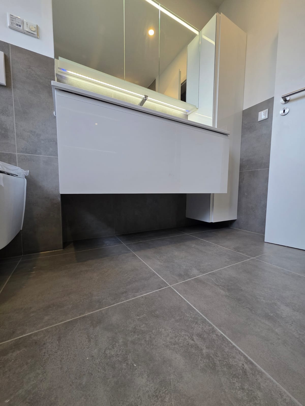 Nowoczesna łazienka po remoncie: szare płytki podłogowe i ścienne, biała szafka pod umywalką z oświetleniem LED oraz lustro. Minimalistyczny styl.