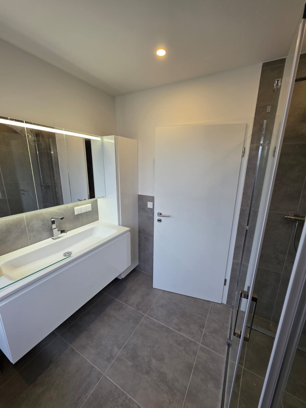 Nowoczesna łazienka z szarymi płytkami, białą szafką z umywalką i lustrem z oświetleniem LED. Kabina prysznicowa z widocznymi okuciami.