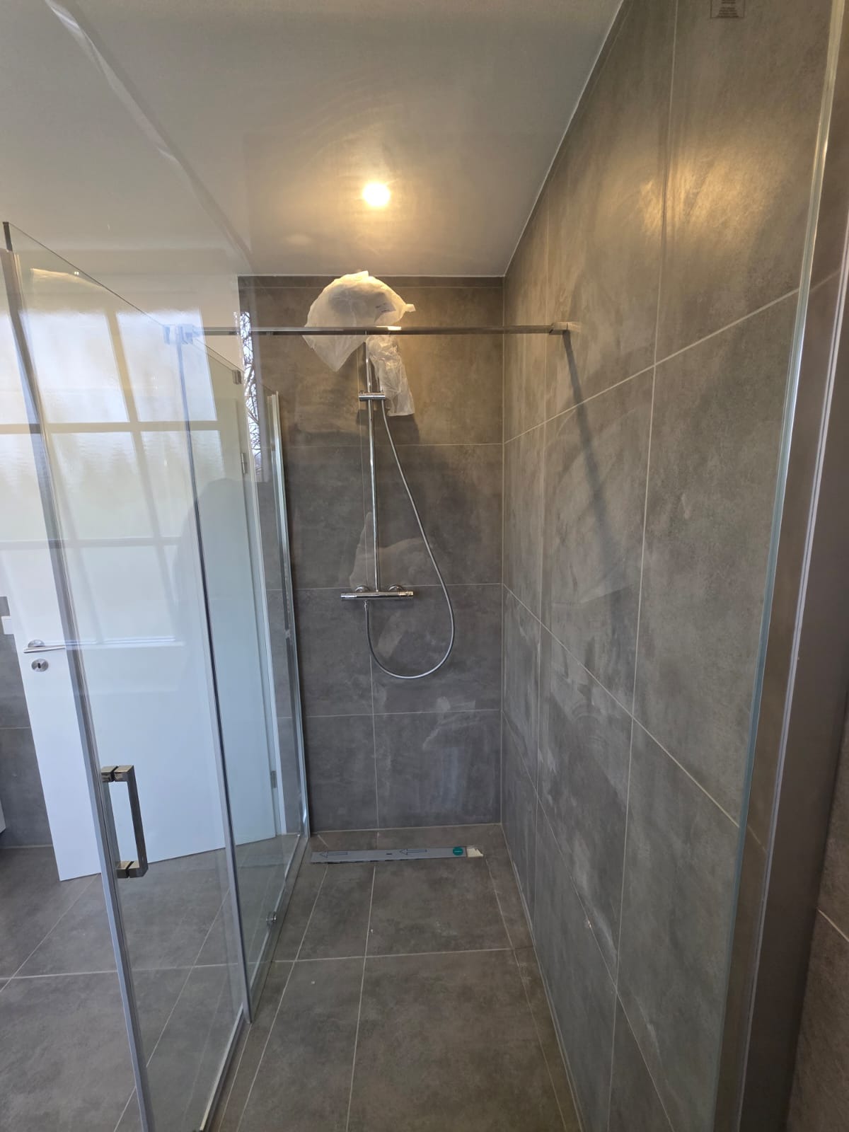 Nowoczesna kabina prysznicowa z szarymi płytkami w dużej łazience. Widoczny chromowany zestaw natryskowy i szklane drzwi kabiny.